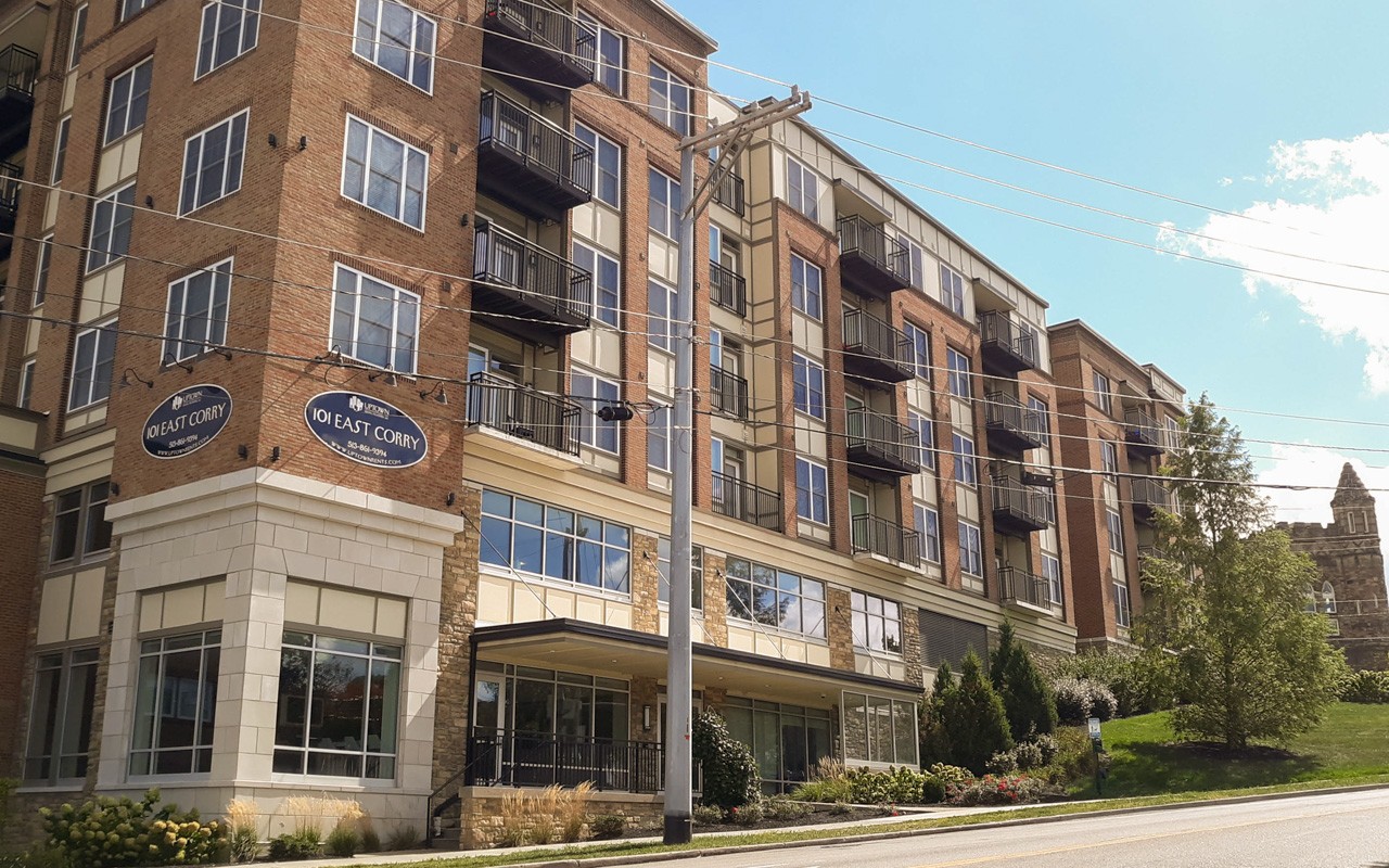 Exterior of 101 East Corry. There are various bushes lining the outside of the building. There is a grass area to the right with a tall tree.