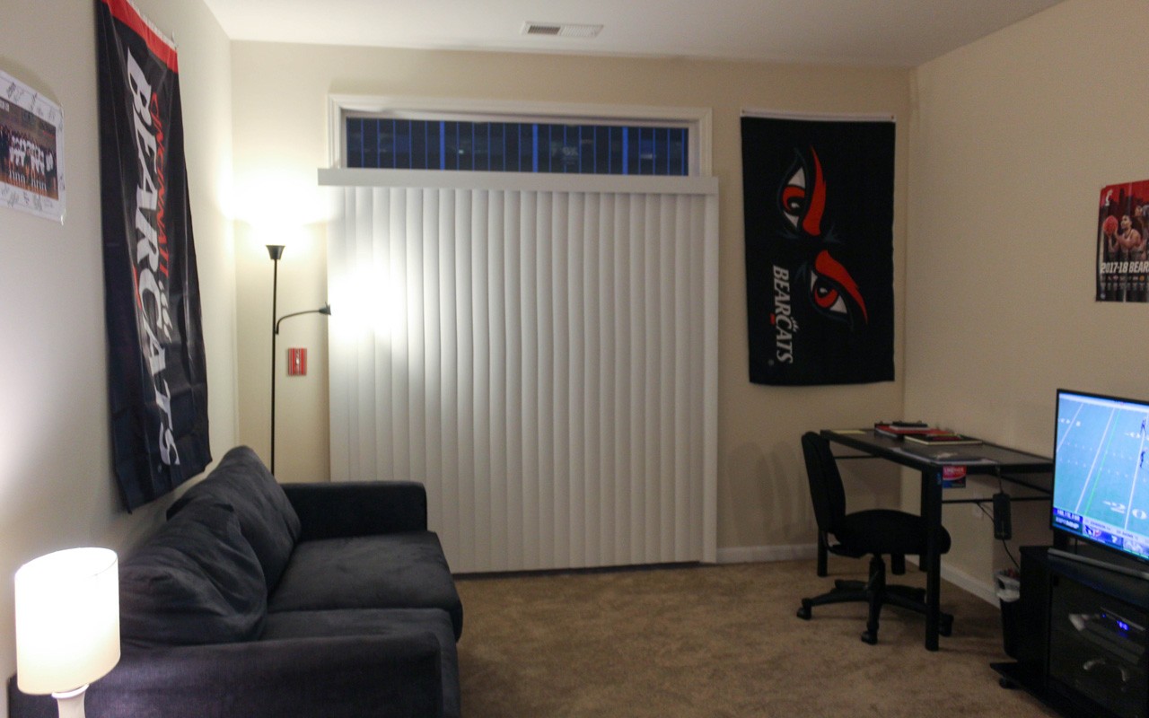 Living room in a U Square apartment with a TV entertainment center and desk,  to the right of the TV is a floor length window with vertical blinds, and there is a couch against the left wall.
