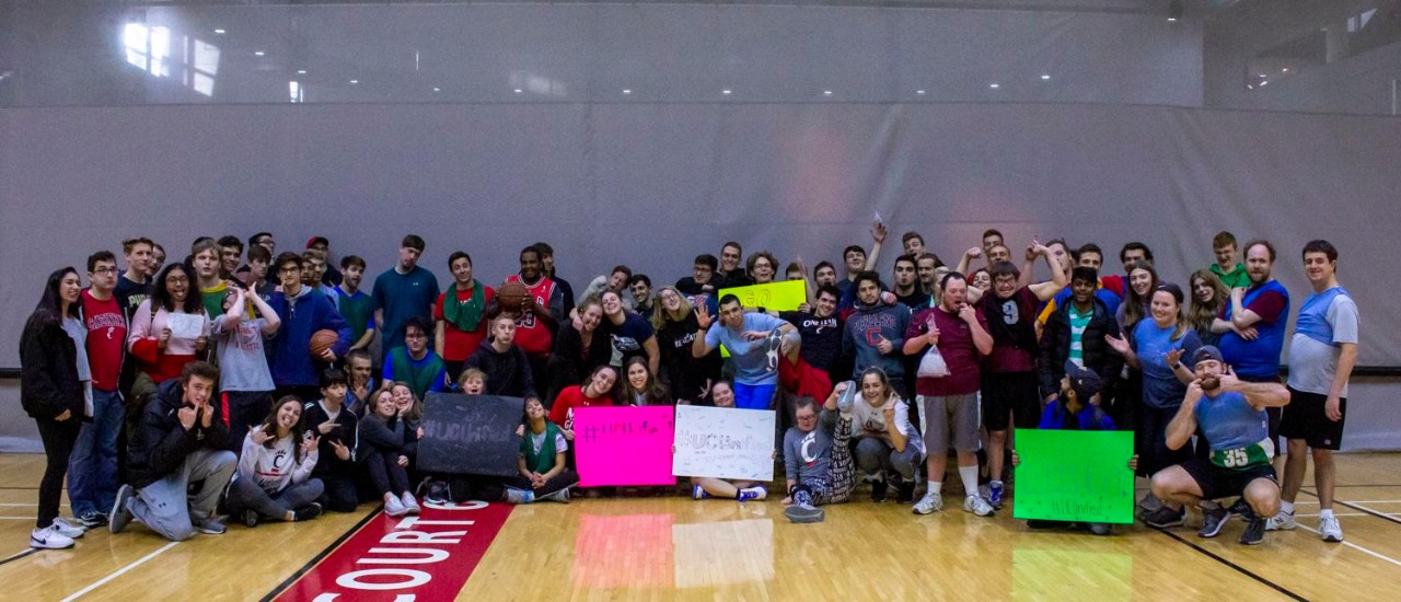 Over 50 student-athletes both from UC Club Sports and the Special Olympics pose for a picture on a basketball court with signs after hosting a successful unified basketball tournament.