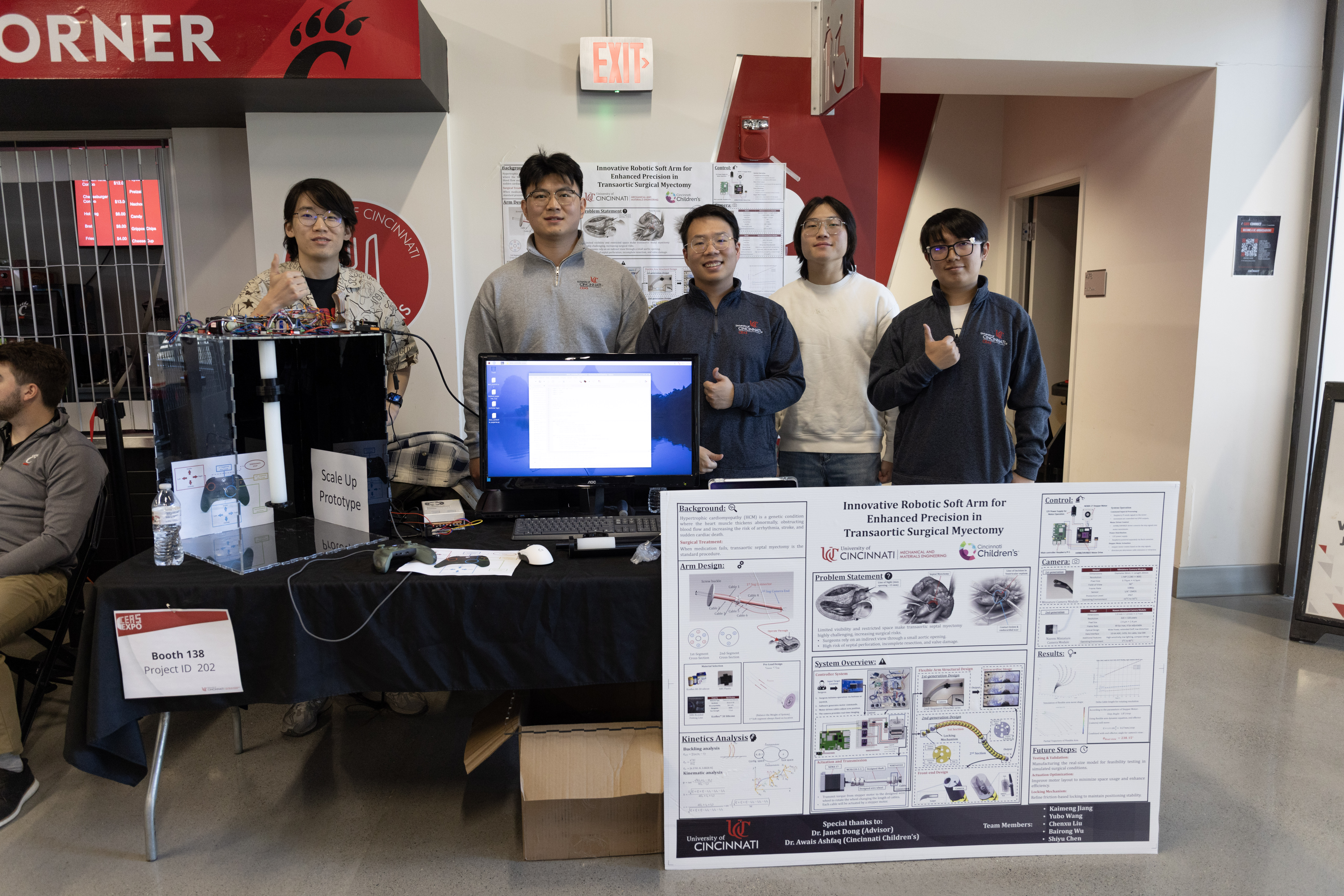 CEAS students stand at their EXPO booth. They give a thumbs up to the camera. a computer monitor is on the table displaying their project information