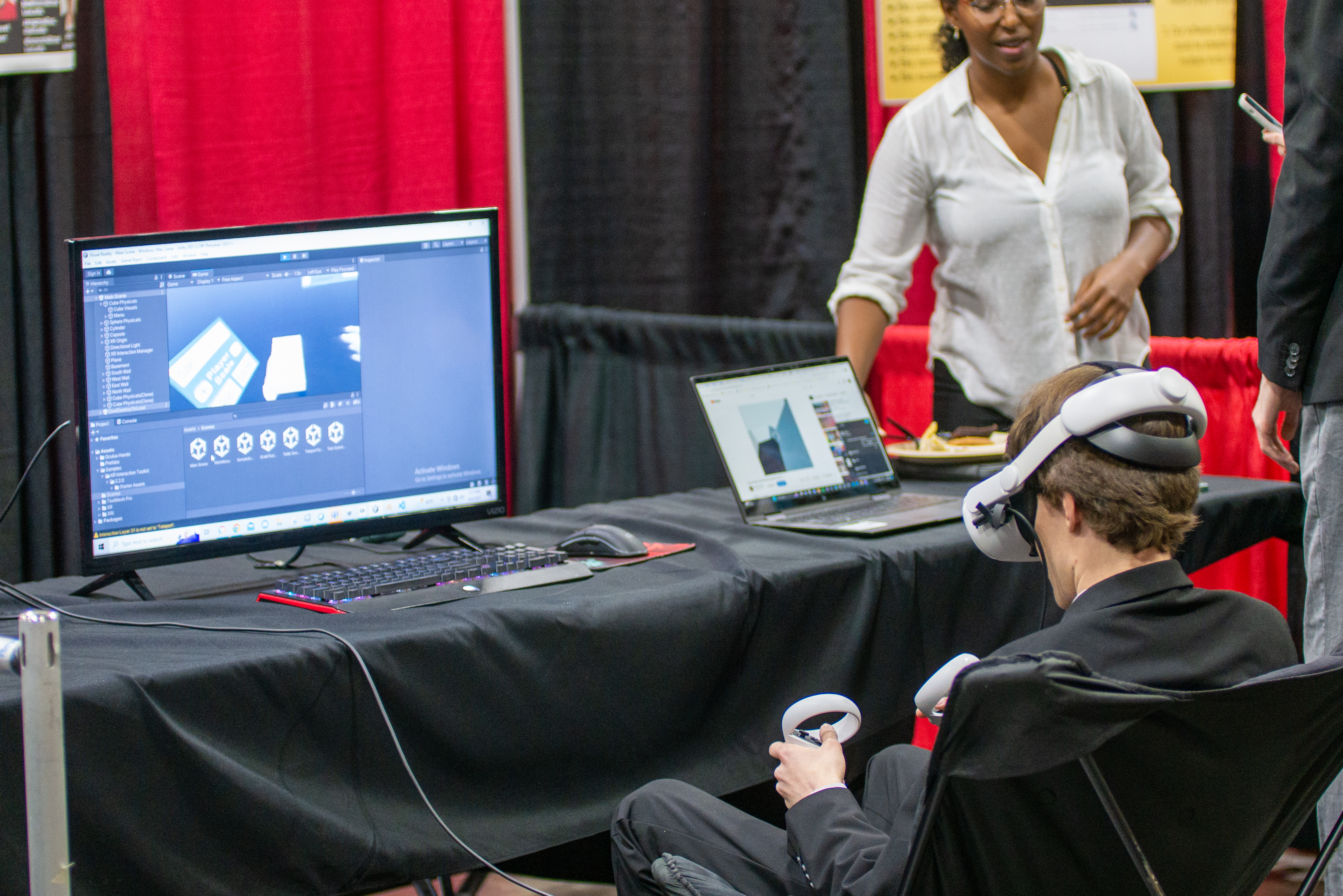 An EXPO attendee sits at a table and wears a VR headset and holds a controller, demo-ing a student project