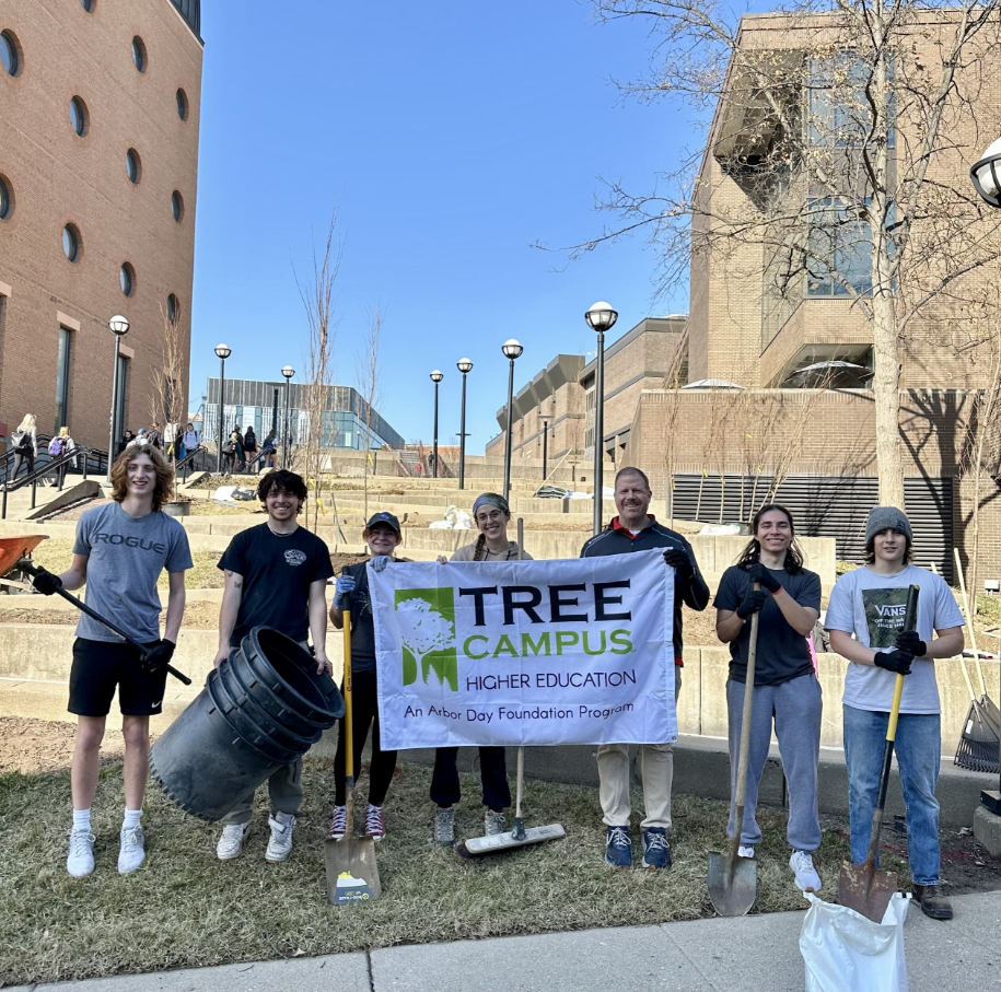 seven students tand with shovels and a banner that says Tree Campus Higher Education near Langsam Library