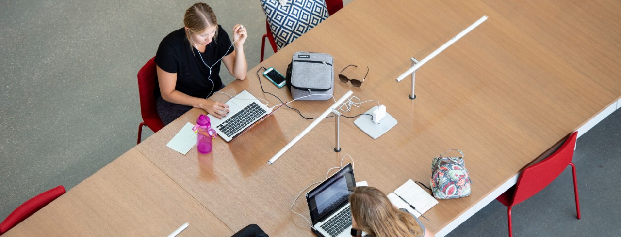 an overhead view of a large table with two students sitting across each other with laptops, backpacks and other study materials.