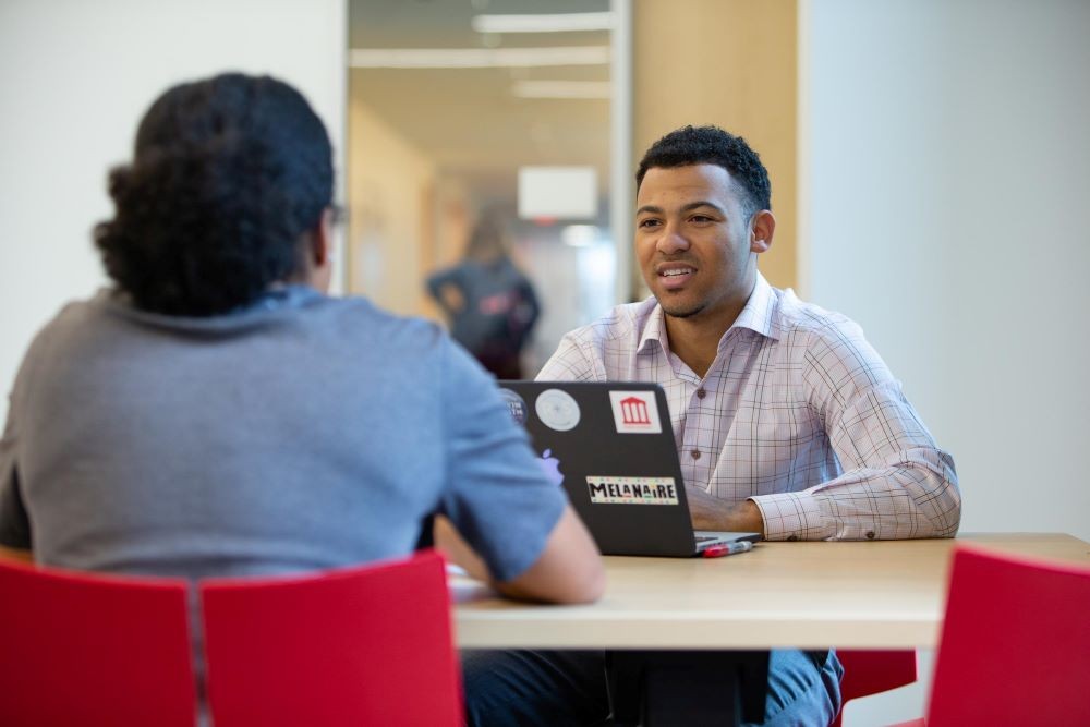 young person in suit sitting in front of a computer looking at someone across a table