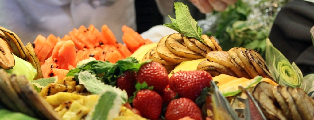 A buffet with various fruits. A food worker's gloved hand is adjusting the display of fruits.