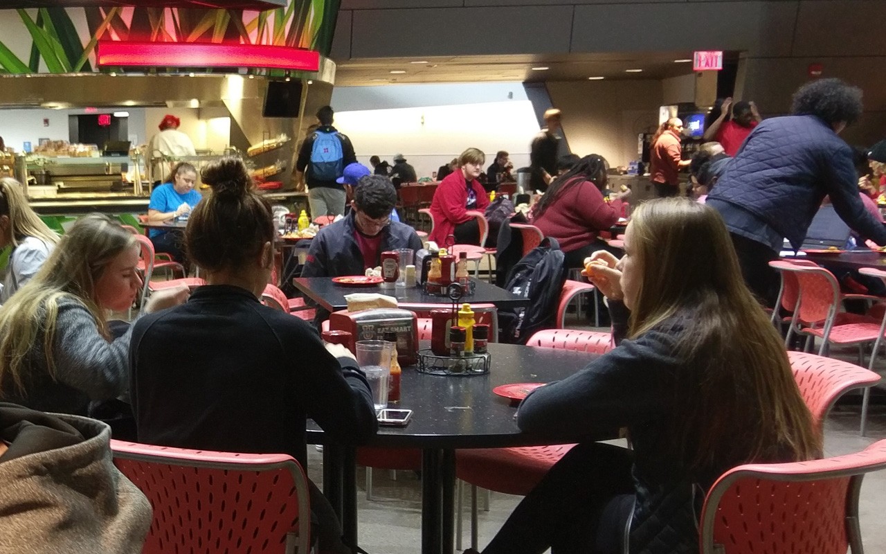 Three students sit eating at a table in Center Court dining center. In the background, are several other table with students sitting and eating as well as the buffet line.