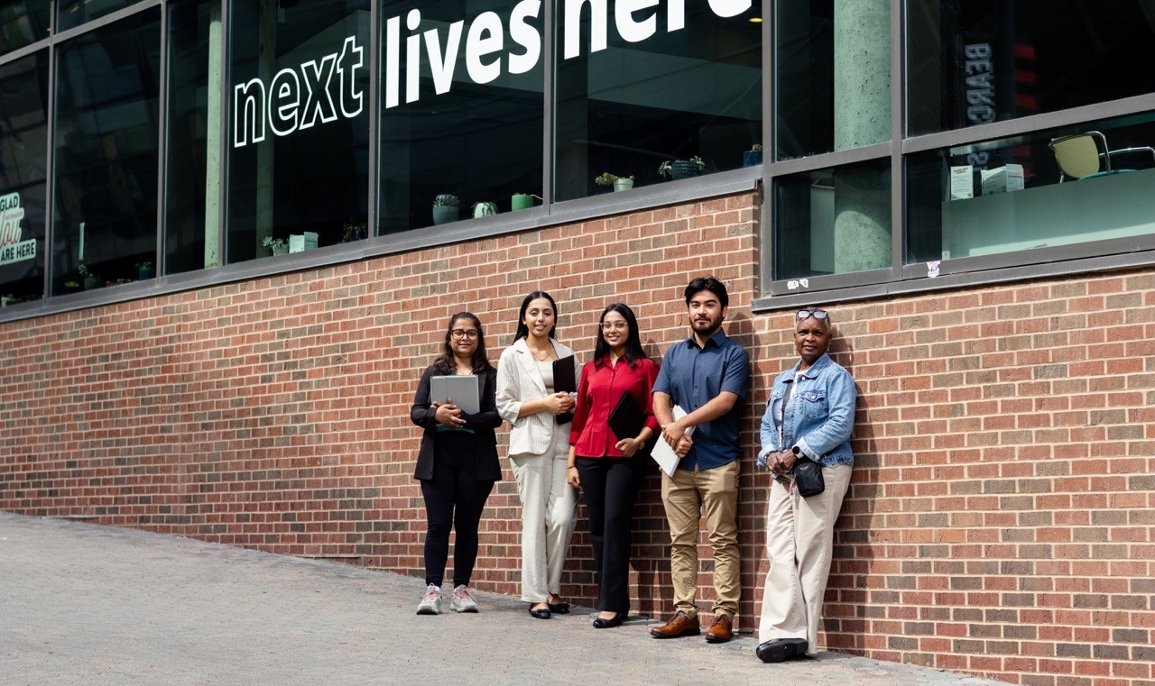 5 students standing in front of the "Next Lives Here" sign on Main Street