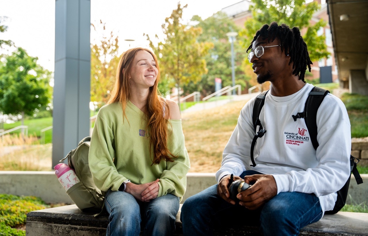 2 students talking and sitting on a bench outside on campus