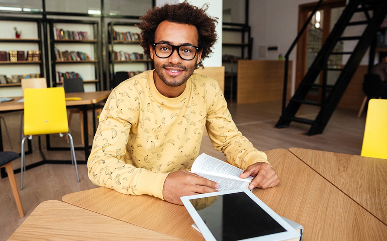Smiling african american young man in glasses with blank screen tablet sitting in library