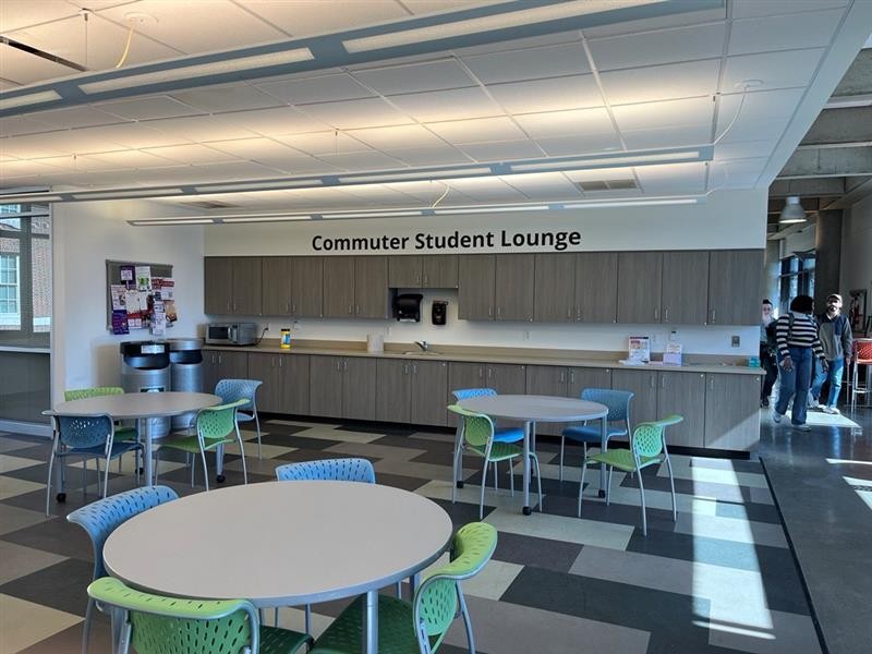 A large open space with a group of three tables with chairs. A long wall features a kitchen with microwave, sink, and supplies. A sign reading Commuter Student Lounge is on the wall.