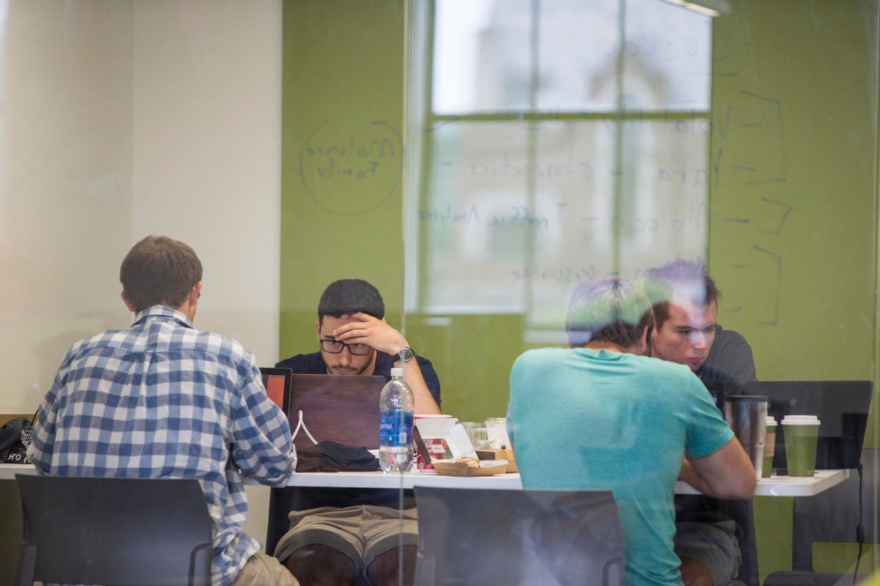 A student reads from a text book, holding a dry erase marker with his back to a math problem on the board. 