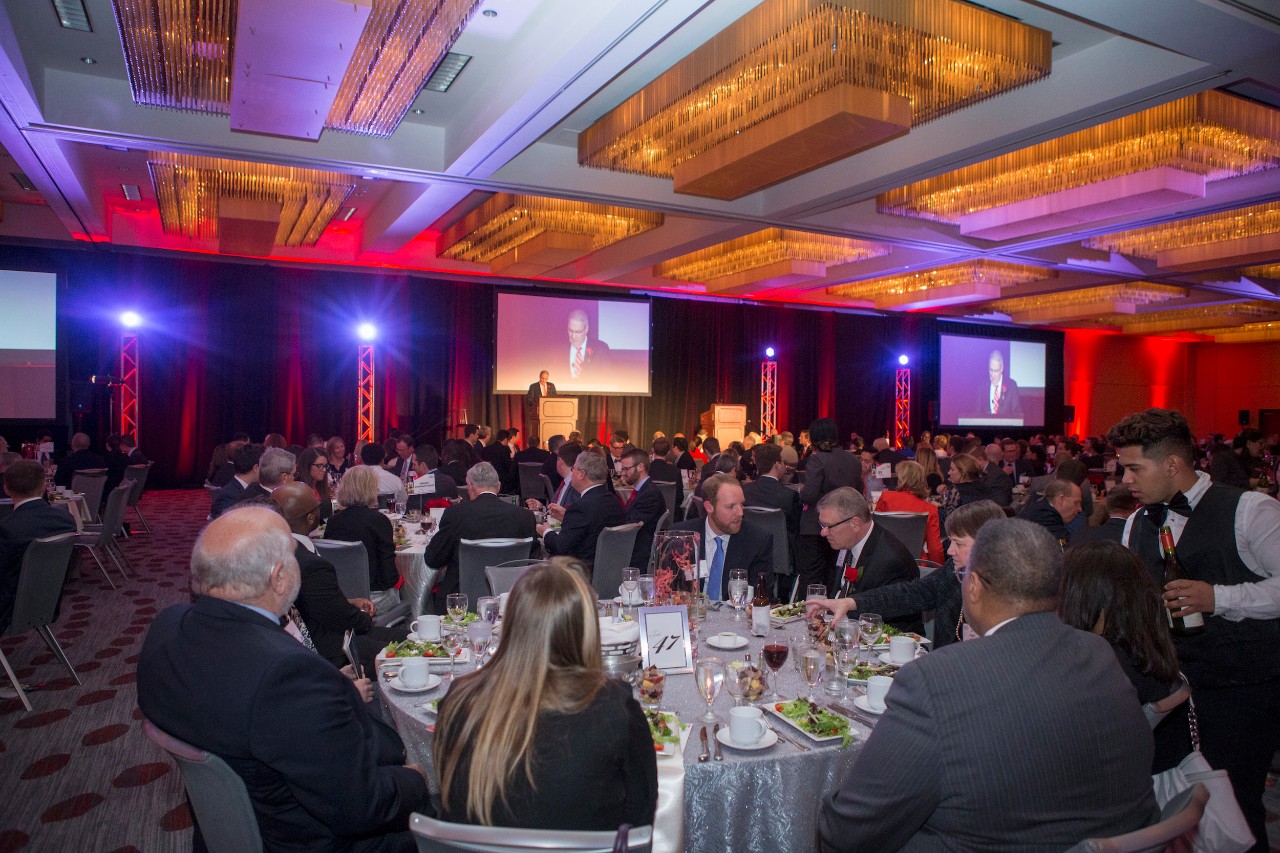 A large banquet hall with people seated at round tables. A speaker at a podium is in the background.