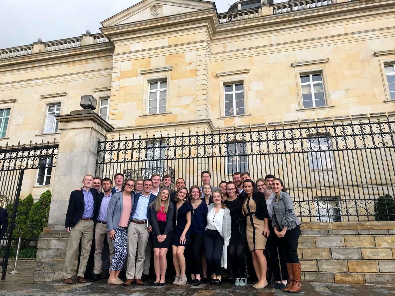 A group of young men and women in nice clothing stand huddled together outside an iron fence to a palace