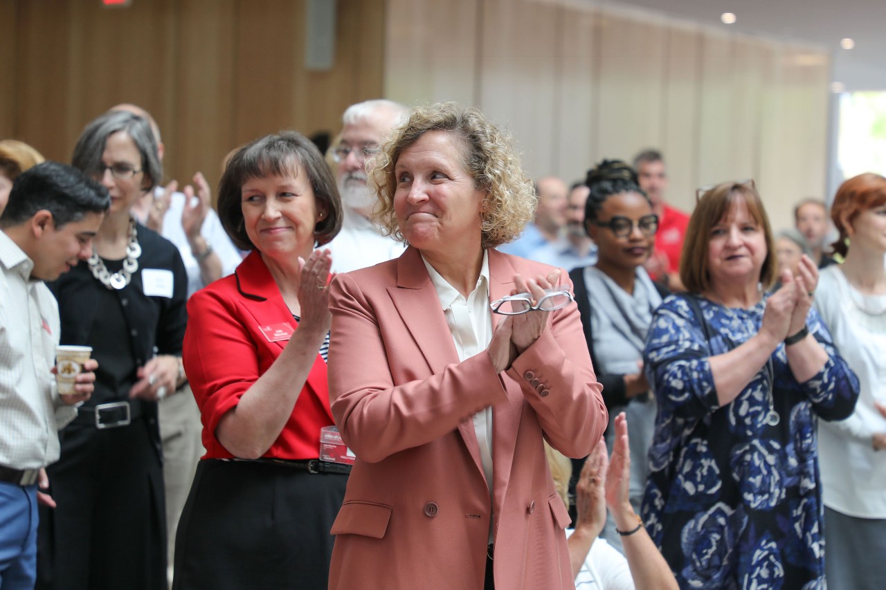 Woman with curly blonde hair in blush-colored blazer stands in foreground with crowd behind her clapping hands