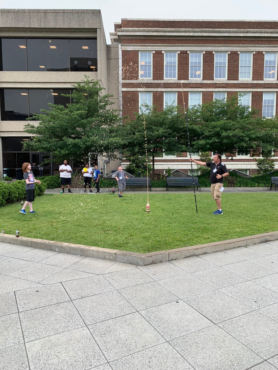 a group of middle school students and parents stand outside a building on a lawn, watching several cola bottles explode with foam