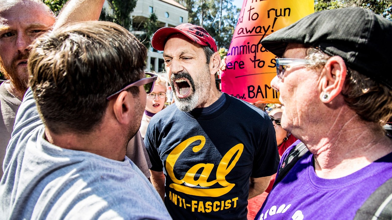 a man screams at two other men among a crowd of protesters