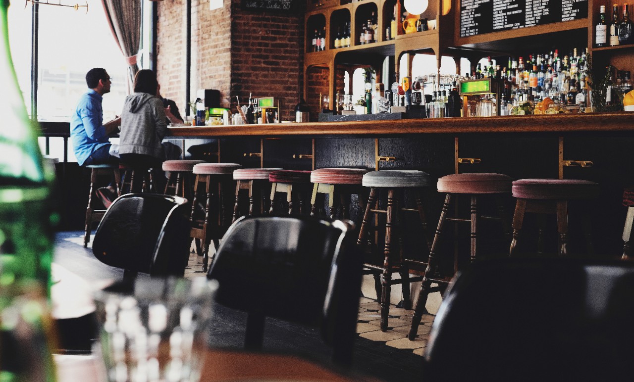 Two people sit on stools at a bar and talk to each other
