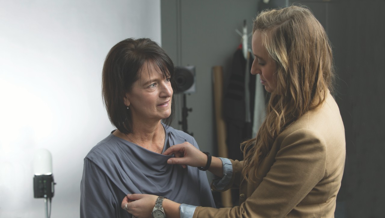 Two women, one of whom is adjusting a garment being worn by the other. 