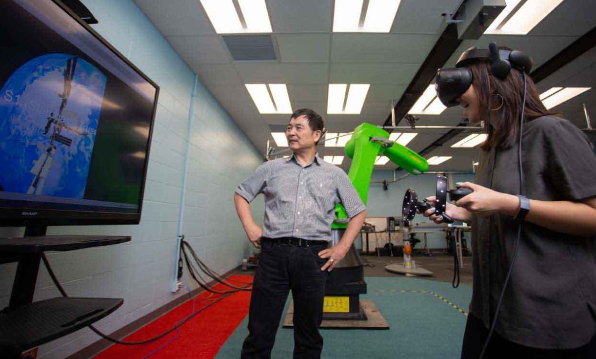 Ou Ma and a student in a VR helmet look at a screen displaying the International Space Station.