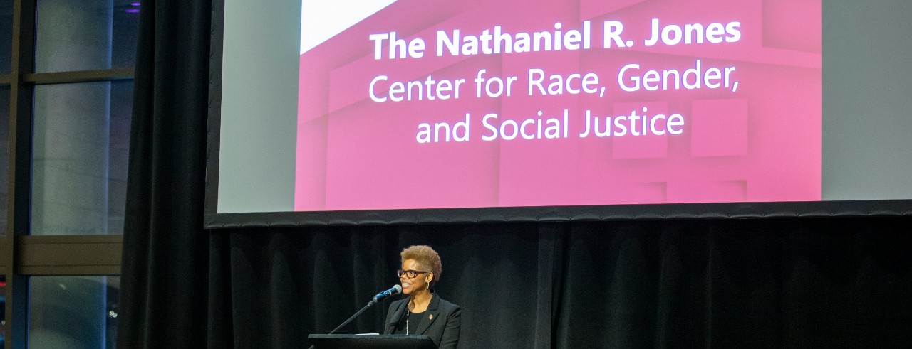 a woman stands at a podium in front of a large red screen
