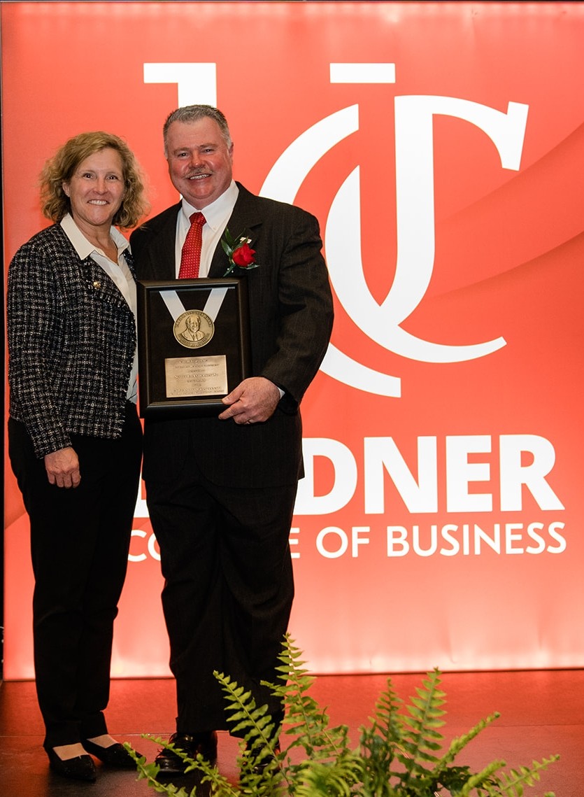 Dean Marianne Lewis stands with award winner Scott Anderson, wearing a dark suit and a red tie and holding a framed medal of his award