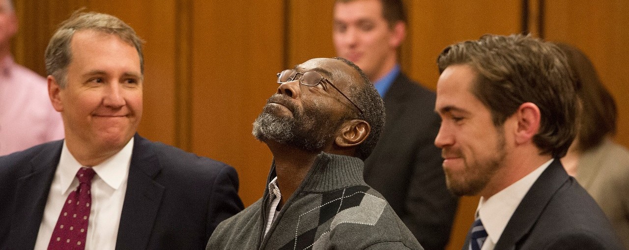 two men in suits surround a man who raises his face upward in joy