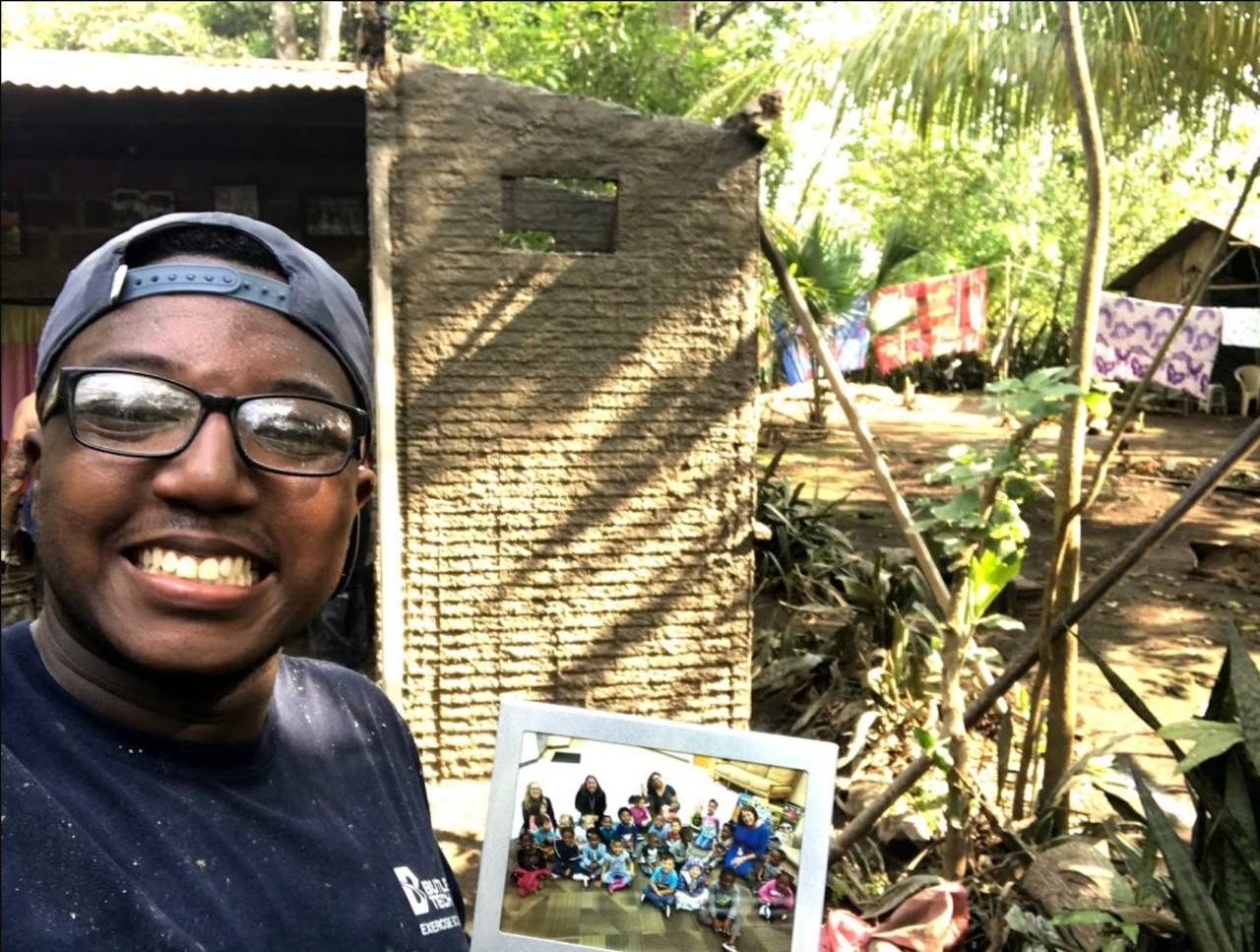 A student holds a photo while standing in front of a hut in a forest