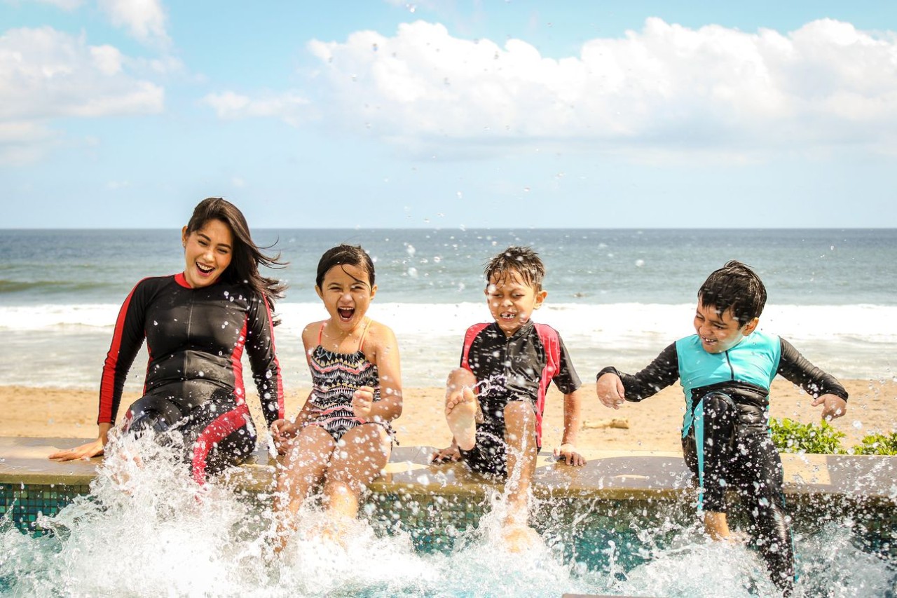 three children and a woman sitting on a ledge by the ocean with water splashing