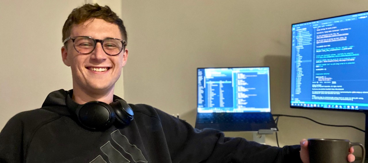 UC student Jakob Wardega sits at two computer screens while looking down at his smartphone.