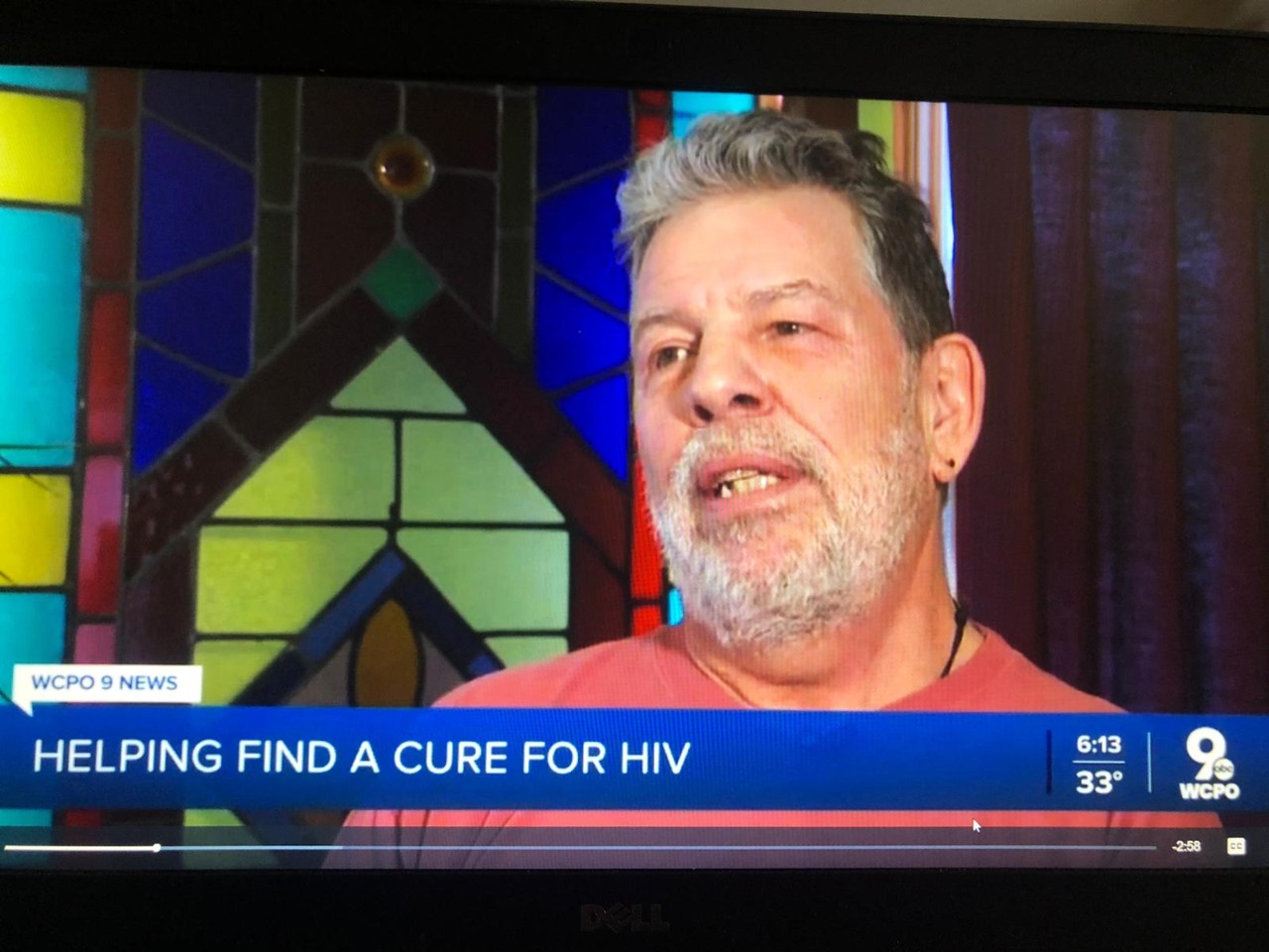 A screen shot from a TV broadcast with a bearded man talking and the headline says helping find a cure for HIV
