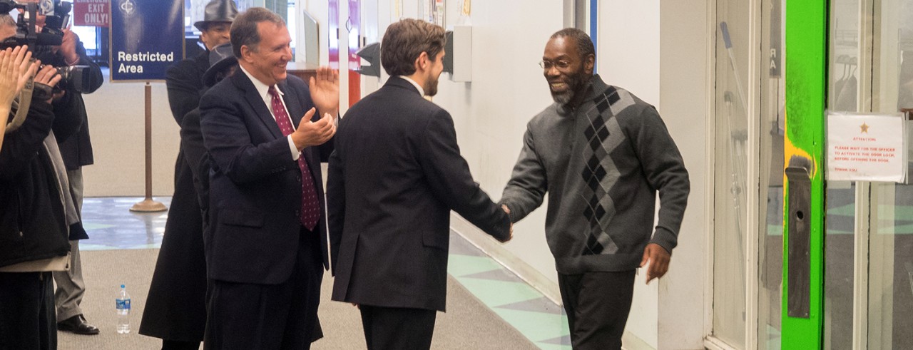 A man shakes the hand of a man in a suit as another man in a suit cheers