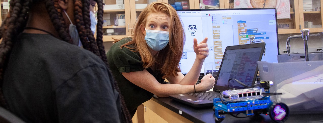 Students in face masks discuss programming a robot on a table in front of them next to an open laptop computer.