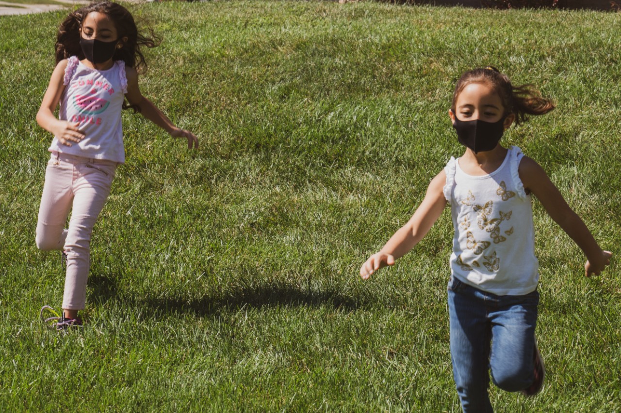 Two children wearing masks run in a grassy field