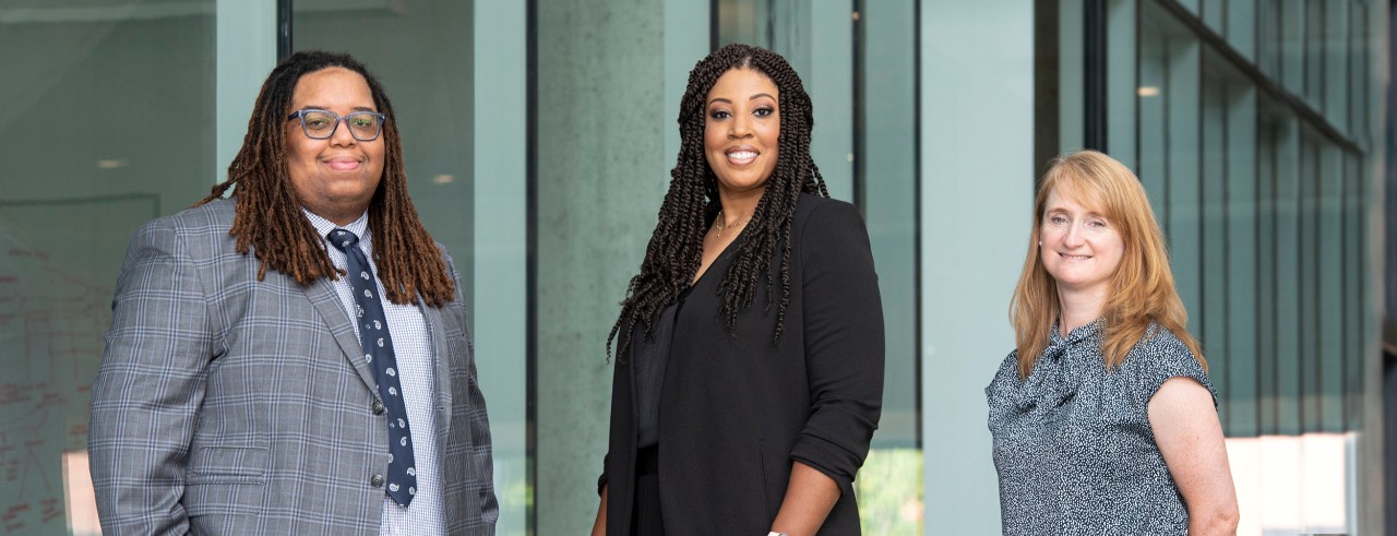 From the left, a man, a woman and another woman pose inside a college building