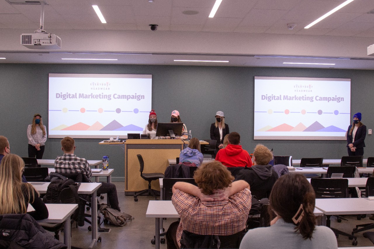 Five students stand in front of two projection screens for their presentation.