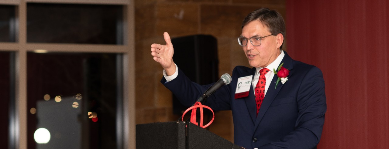 Chris Dobrozsi speaks from a podium and gestures to the unseen audience in front of him