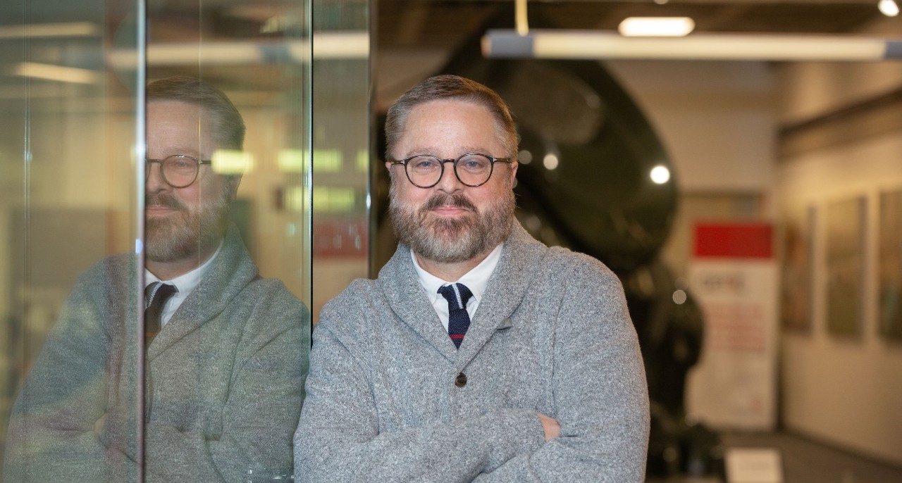 Professor of journalism, Jeffrey Blevins leaning against a wall in the School of Journalism , wearing a grey sweater, white shirt and black tie. 