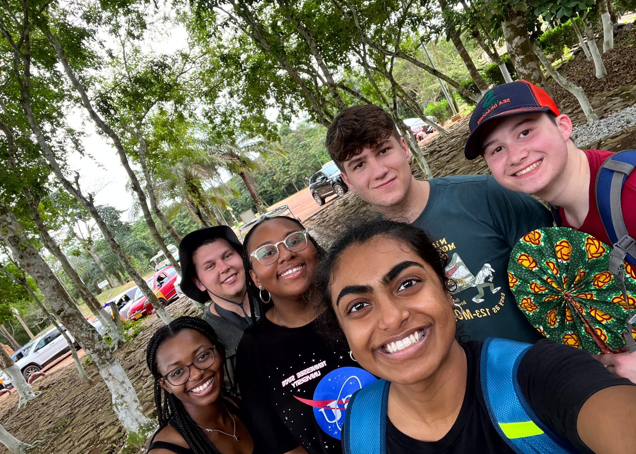 A group of young people smile for a selfie, standing in a grove of white-barked trees; the ground is covered with roots and stones, and a parking lot is visible beyond it