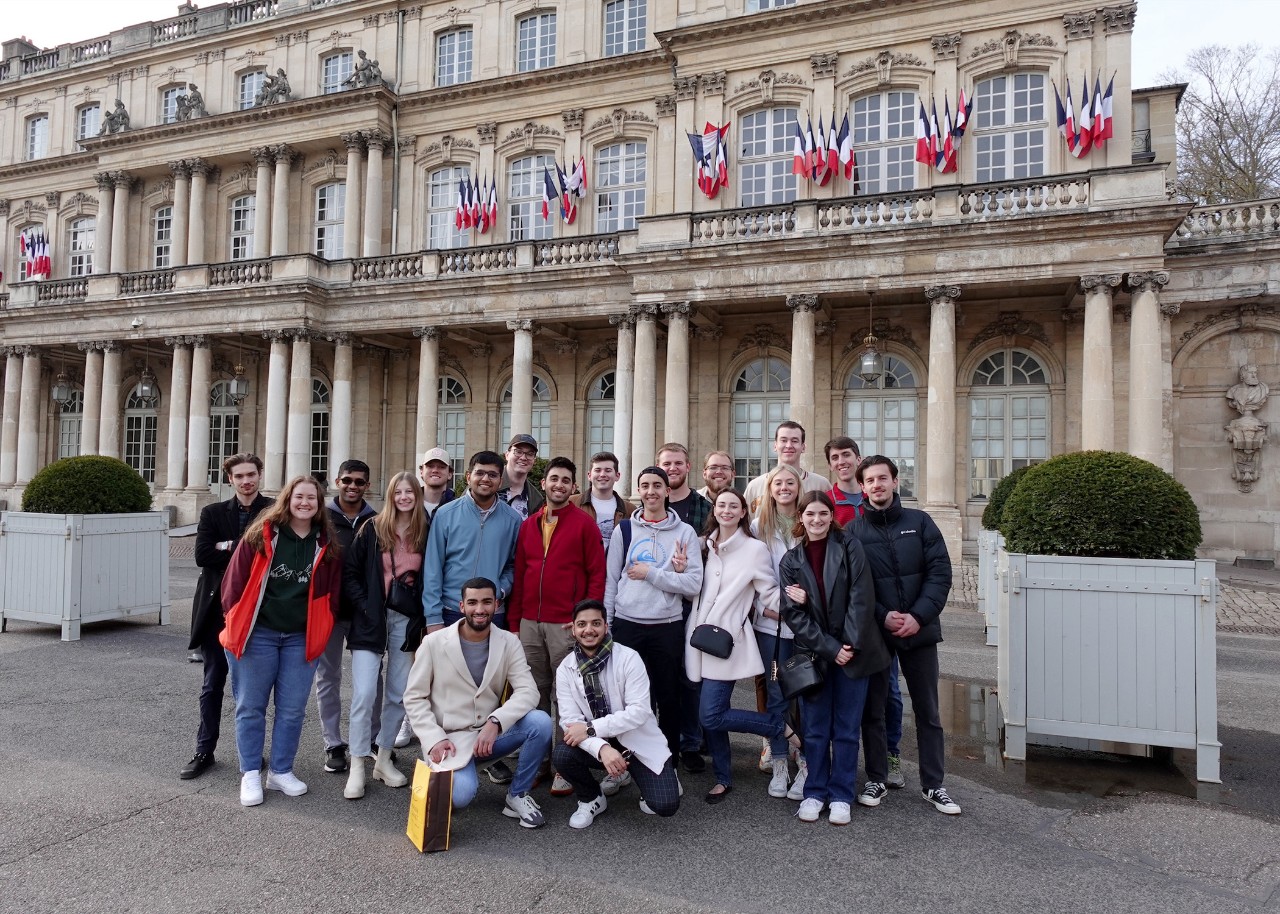 a large group of young people pose for the camera, standing and kneeling in front of a pillared stone building with many arched windows and adorned with bunches of small French flags