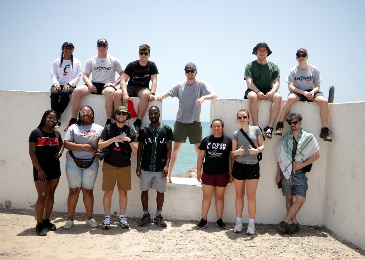 A group of young people pose sitting on a whitewashed wall and standing in and in front of an embrasure in the wall, which is at Elmina Castle in Ghana; behind them, blue sky and blue water beyond a rocky coast