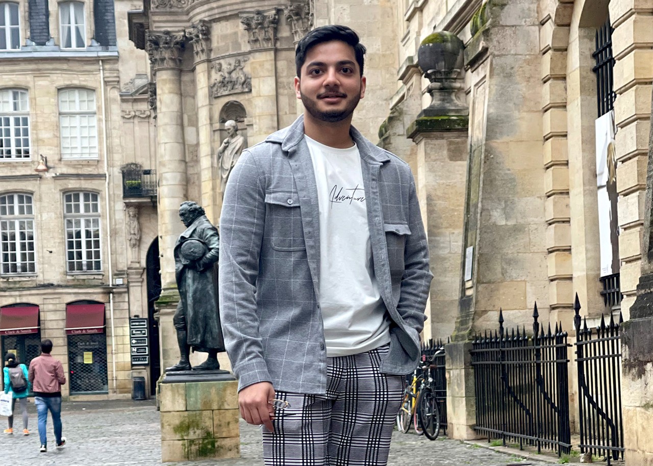 A young man stands in a cobbled street; behind him are stone buildings with arched windows, columns, wrought-iron railings and sculpted friezes, a bronze statue of a man in 18th-century dress stands behind him