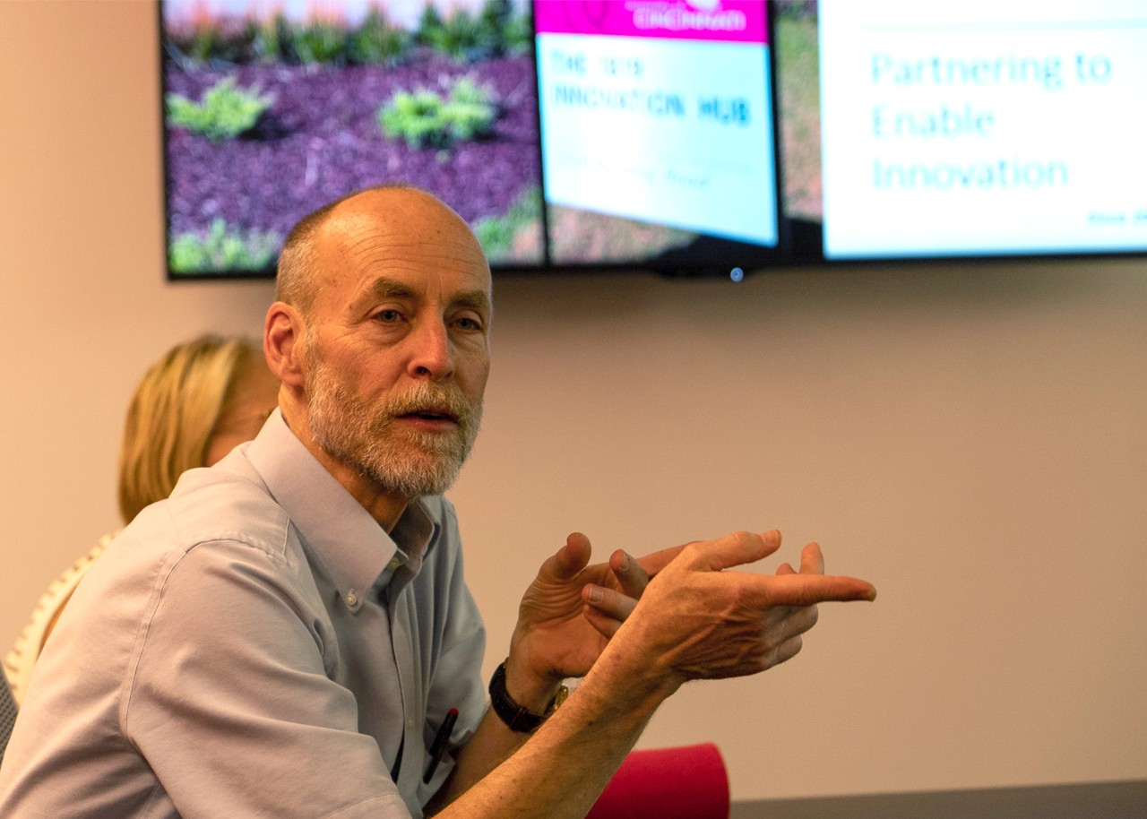 Rutz sits in a room, facing to the right, gesturing in conversation with someone off-camera; behind him on the wall  are 3 digital screens, one of which reads, "partnering to enable innovation"; a woman can also partly be seen, seated behind him