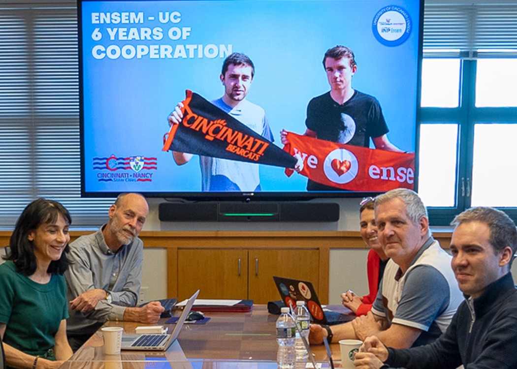 a group is seated around a table in front of a digital screen with a slide that says "ENSEM-UC, 6 years of cooperation" and shows two people holding school pennants