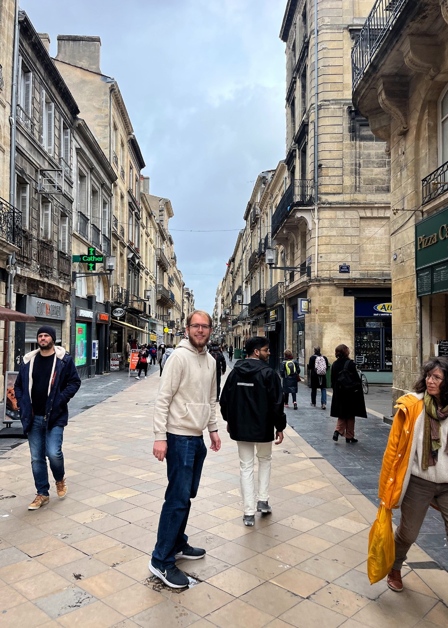 a young man turns to look toward the camera on a pedestrian street; buildings with wrought-iron balcony railings and shopfronts on either side of the street converge toward a central perspective point
