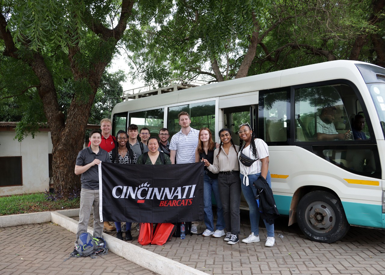 A group of people stand beside a small bus, holding a banner that reads "Cincinnati Bearcats"