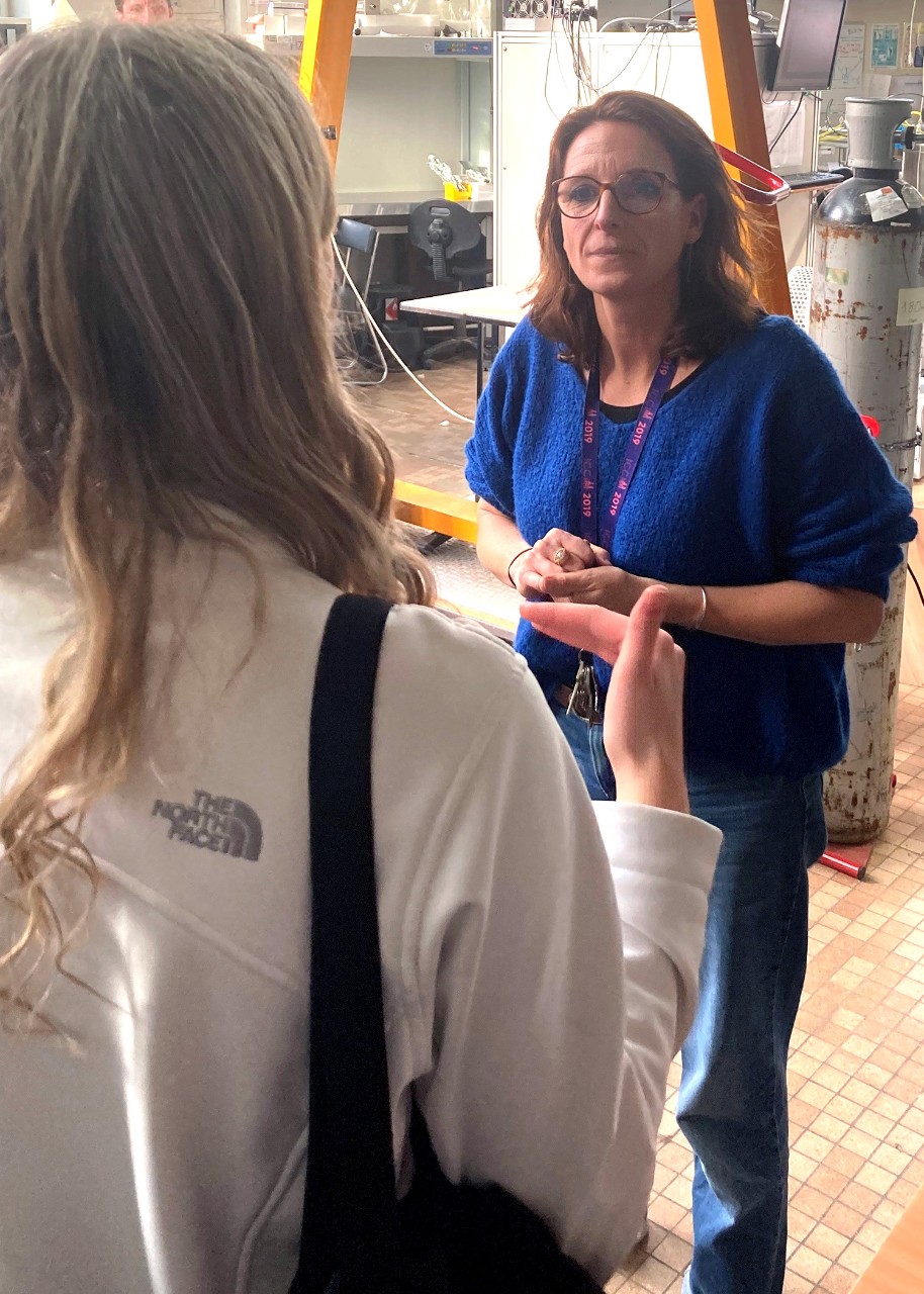 A young woman stands with her back to the camera in a lab space full of mechanical and digital equipment and wooden frames; facing her, a woman in jeans and a blue pullover is listening to her intently, her hands clasped in front of her