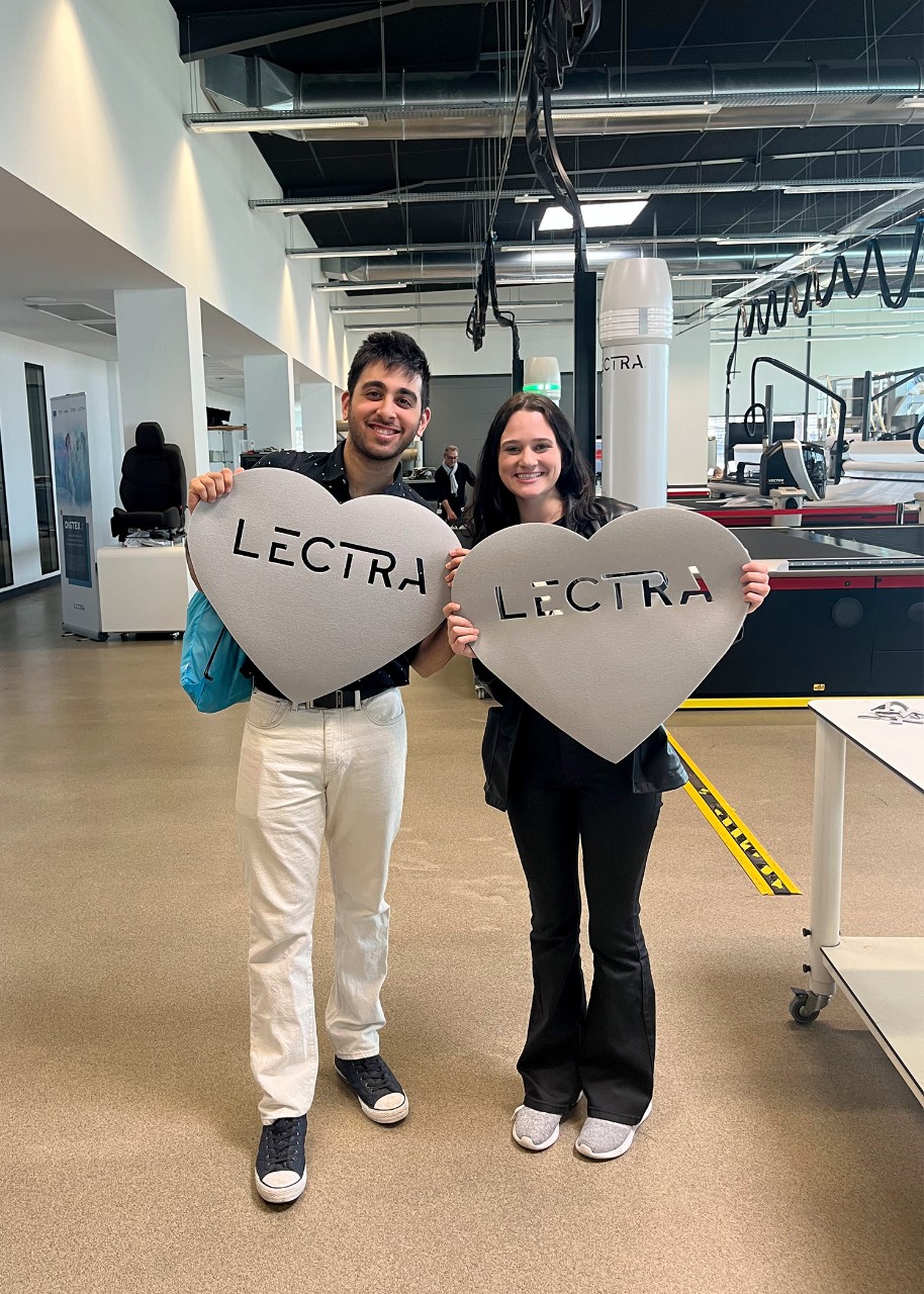 Two students pose, smiling, side by side, on a factory floor; they are holding large hearts cut from sheets of plastic with the word “Lectra” laser cut into them; high-tech machinery is visible behind them, while electric cables drape from the ceiling