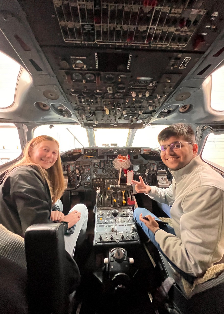 Two people sit in the cockpit of a flight simulator, turning to smile at the photographer behind them