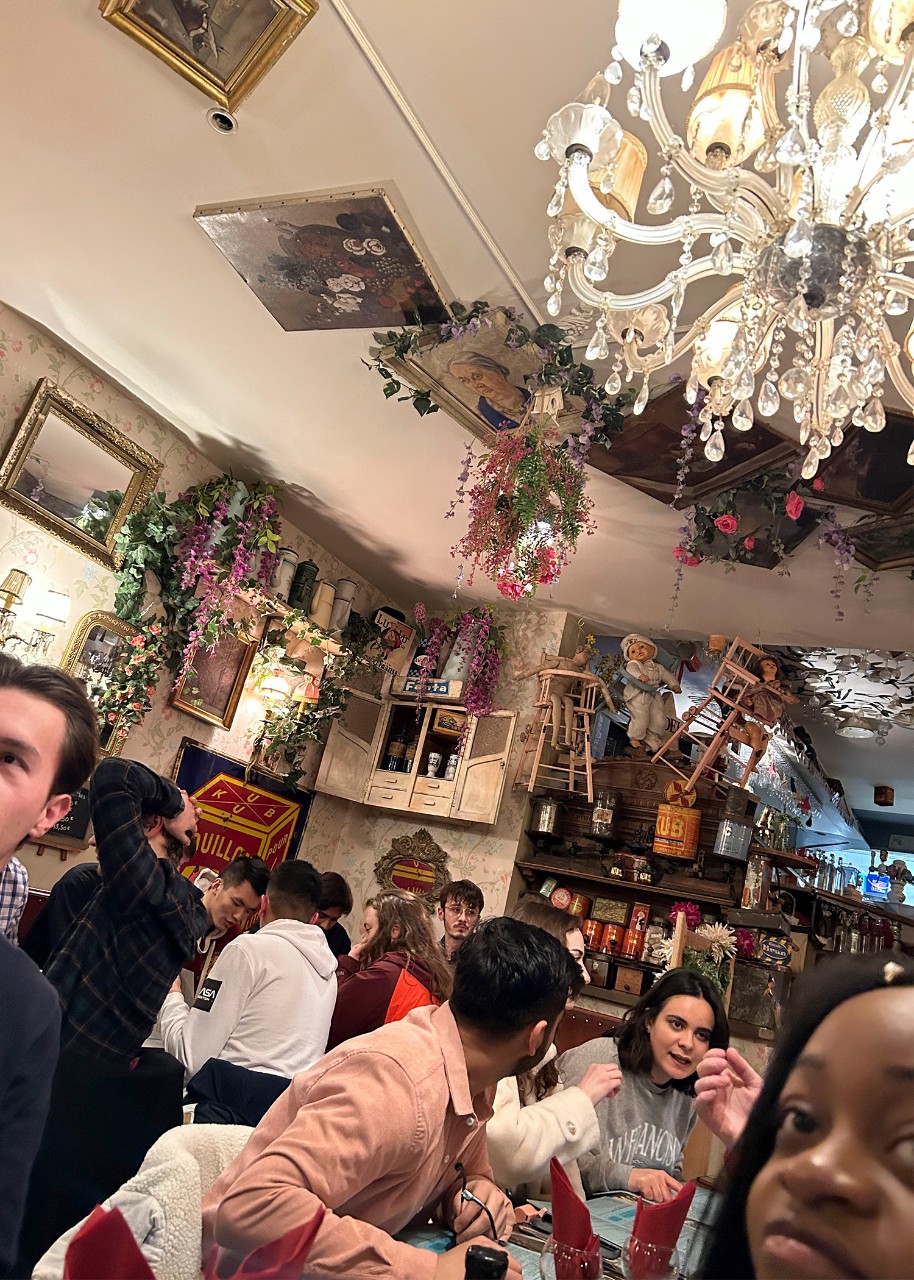 A crowded restaurant scene shot from a skewed perspective shows groups talking at tables, with a crystal chandelier, bunches of flowers, and painted canvases on the ceiling above them; beyond them is the bar back; in a corner of the foreground, one young woman looks at the camera 