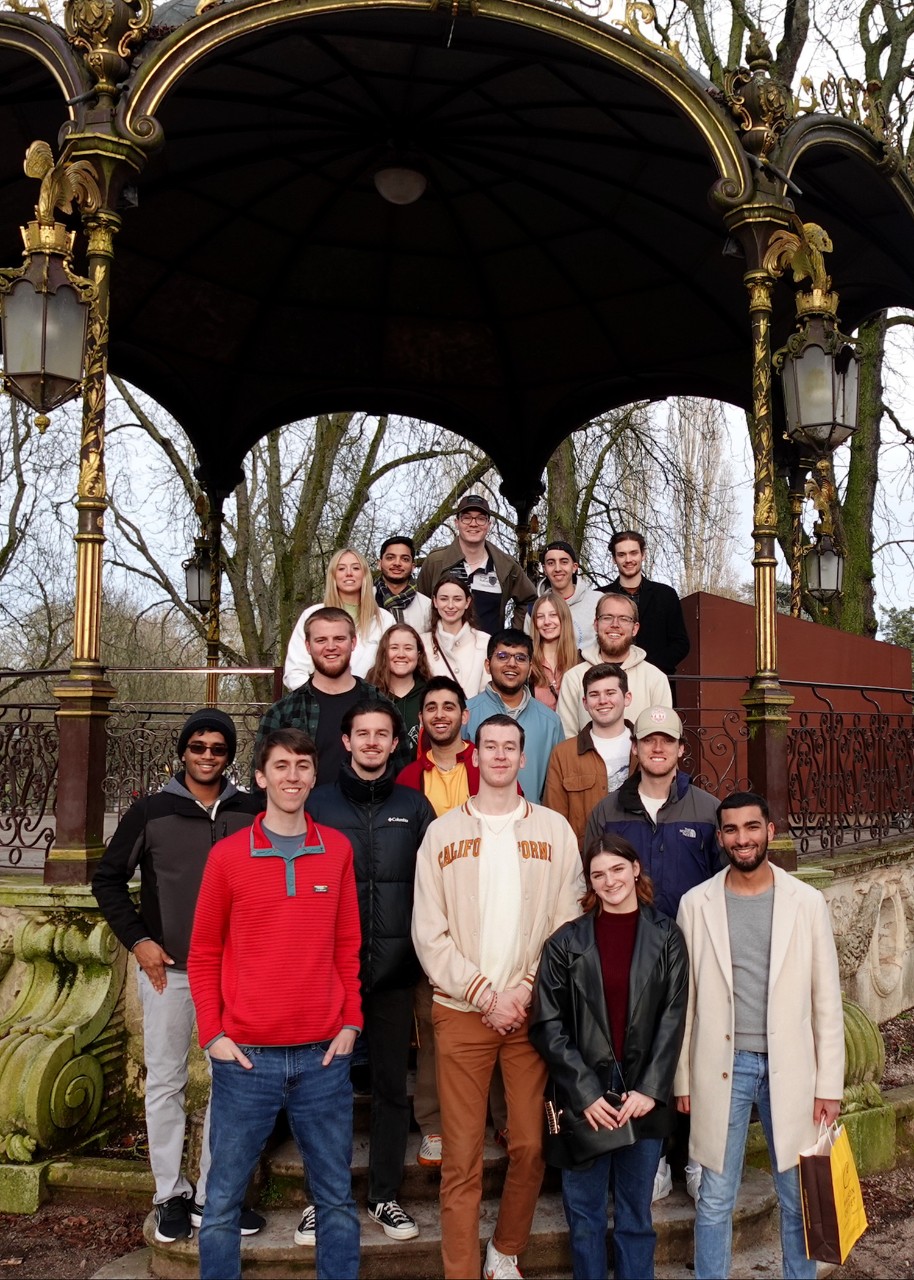 A group of students pose for the camera on the steps of a pavilion in a park; the pavilion has a roof with multiple arches, a wrought-iron railing, gilded columns and hanging lanterns